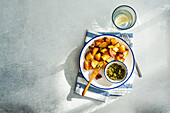 Knusprig gebratene Kartoffeln neben einer Schüssel mit grünem Pesto auf einem blau gestreiften Teller, über einer blau gestreiften Serviette, mit einem Glas Wasser mit Zitronenscheibe im Hintergrund, auf einer strukturierten Oberfläche bei Tageslicht
