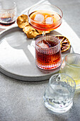A curated selection of alcoholic beverages artfully arranged on a concrete surface, featuring glasses with cognac and cinnamon, cherry liqueur with ice, and other drinks accompanied by dried citrus slices
