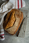 Top view of freshly baked loaf of rye sourdough bread sits cooling on a wire rack, accompanied by a bread knife and a white cloth with red stripes