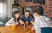 Eine Mutter mit ihren Kindern, die lächelnd ein Würfelspiel am Familientisch mit Snacks und Getränken während eines gemütlichen Wochenendes zu Hause spielen