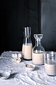 Two glass bottles of homemade oat milk with a glass half full and scattered oats on cloth