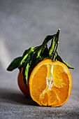 A vibrant, freshly sliced orange with lush green leaves set against a textured grey background, highlighting the fruit's juiciness and natural beauty