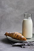 A homemade pastry cone filled with jam and sprinkled with powdered sugar, placed beside a full milk bottle on a ceramic plate, set on a textured grey background