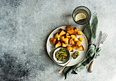 Top view of golden roasted potatoes on a white plate with green pesto in a small bowl, a glass of water, and cutlery wrapped in a green napkin on a textured grey background