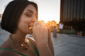 A young woman bites into a delicious burger against the backdrop of Madrid's iconic Cuatro Torres Business Area during sunset.