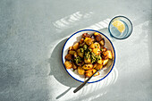 Draufsicht auf sonnenbeschienene Bratkartoffeln mit einem Klecks grünem Pesto auf einem weißen Teller mit blauem Rand, daneben ein Glas Wasser mit Zitrone, das einen Schatten auf eine strukturierte graue Oberfläche wirft