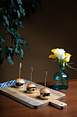 Elegante Präsentation von Mini-Burgern aus Rindfleisch, überbacken mit geschmolzenem Cheddar-Käse, serviert auf einem Holzbrett mit einer dekorativen Vase mit Rosen.