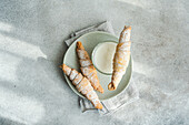 Top view of homemade pastry cones filled with jam and sprinkled with powdered sugar, alongside a glass of milk on a ceramic plate, set on a textured grey surface
