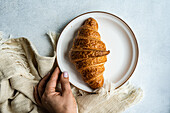 Top view of anonymous person hand holding a plate with a freshly baked croissant on a textured cloth, capturing a simple and cozy breakfast moment