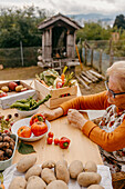 A senior woman examines tomatoes while surrounded by freshly harvested vegetables on a rustic farm table with a garden shed in the background