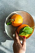 Anonymous hand with red nail polish selects a ripe orange from a metallic bowl under the sunlight, creating shadows