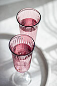 Zwei rosafarbene Gläser werfen von oben zarte Schatten auf einen weißen Tisch mit natürlichem Licht. Generatives AI-Bild
