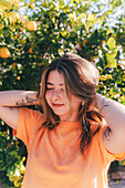A smiling young woman in a vibrant orange t-shirt basking in the sunlight amidst lemon trees.
