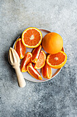 From above sliced and whole oranges arranged on a ceramic plate next to a wooden citrus juicer on a textured gray surface.