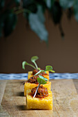 Bite-sized polenta canapé topped with a savory tomato and onion stew, garnished with microgreens, presented on a rustic wooden surface.