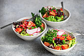 Three bowls of vibrant vegetable salad with lettuce, arugula, radishes, cherry tomatoes, green onions, and pomegranate seeds on a grey countertop