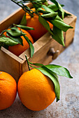 Reife Orangen mit frischen grünen Blättern werden ausgestellt, einige liegen auf einer strukturierten Oberfläche, andere sind in einer Holzkiste eingebettet und vermitteln den Eindruck einer frischen Ernte