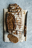 Top view of loaf of rye sourdough bread, partially sliced, with a bread knife on a beige kitchen towel against a textured backdrop
