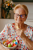 A content elderly lady holds a bowl of vibrant candies while eating a biscuit, showcasing delightful small pleasures at an advanced age