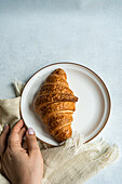 Top view of anonymous person hand holding a plate with a freshly baked croissant on a textured cloth, capturing a simple and cozy breakfast moment