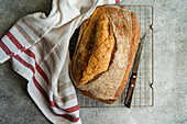 Top view of freshly baked loaf of rye sourdough bread sits cooling on a wire rack, accompanied by a bread knife and a white cloth with red stripes