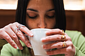 Eine junge Frau mit dunklem Haar und zierlichen Ringen nippt an einer weißen Kaffeetasse, ein intimer Moment, der in einem gemütlichen Innenraum eingefangen wurde.