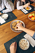 Close-up of anonymous hands at a family breakfast table with a selection of cookies, muffins, cereal with milk, and a fruit bowl