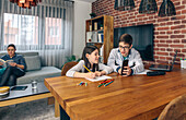 Children engage in drawing and using a smartphone while a woman reads a book on the couch, depicting a relaxed family weekend