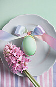 Draufsicht auf ein weiches, pastellfarbenes Ostergeschirr mit einem dekorierten Ei, das mit einem bunten Band auf einem weißen Teller gebunden ist, begleitet von rosa Hyazinthenblüten.