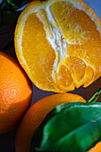 A juicy, freshly sliced orange is in sharp focus with whole oranges and leaves in the background, set against a blue surface
