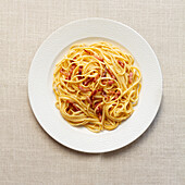 Draufsicht auf traditionelle Spaghetti Carbonara auf einem strukturierten weißen Teller vor einem neutralen Tischtuch.