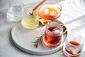 A stylish selection of alcoholic drinks in various glasses, accented with a cinnamon stick, rosemary, and ice, set upon a round concrete tray in soft light