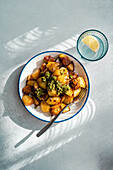 Top view of sunlit roasted potatoes with a dollop of green pesto on a white plate with blue trim, next to a glass of water with lemon, casting a shadow on a textured grey surface