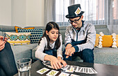 Siblings concentrate on a card game on the living room table, with a boy in a top hat arranging the hand and a girl selecting a card