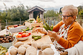 Eine ältere Frau begutachtet sorgfältig grüne Chilischoten an einem Tisch mit einer Auswahl an frischem Gartengemüse