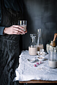 Anonymous person holding a glass of fresh oat milk with ingredients and kitchenware on a dark, textured background