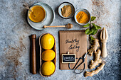 An array of fresh cooking ingredients including lemons, honey, ginger, and spices, alongside a notebook labeled Healthy Cooking on a textured tabletop