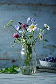 Wildblumen im Einweckglas vor blauem Holzhintergrund, Blaubeeren im Hintergrund