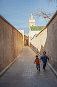 Kinder rennen durch eine alte Gasse vor der Kulisse eines historischen Minaretts in Fez.