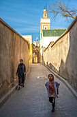 Ein Mädchen und seine Mutter schlendern durch eine sonnenbeschienene Gasse in der Medina von Fez.