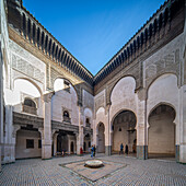 Besucher im verschnörkelten Innenhof der Madrasa Cherratine unter strahlend blauem Himmel.