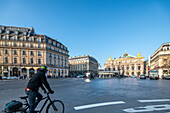 A cyclist crosses an intersection at Pariss Opera Square under a clear blue sky.