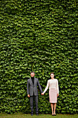 Couple Holding Hands Standing in front of Green Leafy Hedge