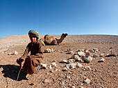 Oman, Ash Charqiya Region, ein junger Beduine in traditioneller Kleidung, Dishdasha und Turban, posiert inmitten eines sehr trockenen, steinigen Plateaus vor einem liegenden Kamel