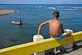 Cuba,Baracoa,a young boy shirtless  looks at fishermen in their boat from the parapet of the road