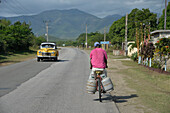 Cuba,a man on a bike is carrying 2 bottle of gaz on a road where a vintage american car from the 50