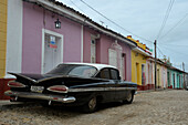 Cuba,Trinidad,an old black American car of the 50s is parked in a paved street in front of low houses with bright colors