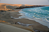 Sultanat Oman,Ostküste,Indischer Ozean,Ein einsamer Mann steht bei Sonnenuntergang oberhalb einer Bucht auf einer ockerfarbenen Sanddüne