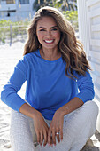 Woman in blue jumper and white trousers sitting on beach