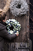 Frau hängt Weihnachtskränze aus Kunststoffkugeln und Naturmaterialien an Holzwand